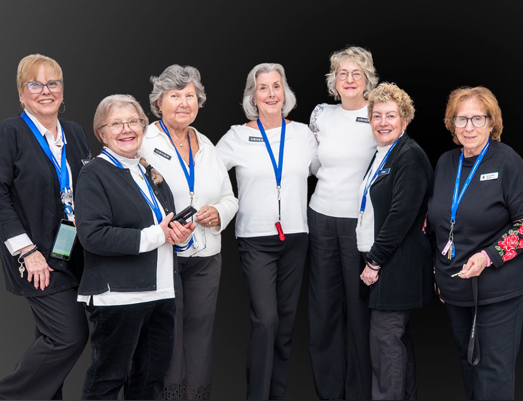 Seven women standing together, all wearing black and white outfits with blue lanyards, against a gray background.