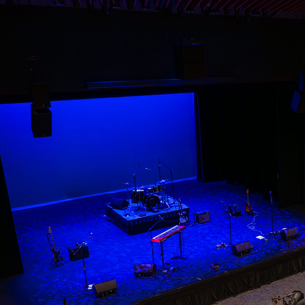 A dimly lit stage with a blue backdrop, featuring technical equipment including a drum kit on a raised platform, a red keyboard, an electric guitar, and microphones, all prepared for a performance.