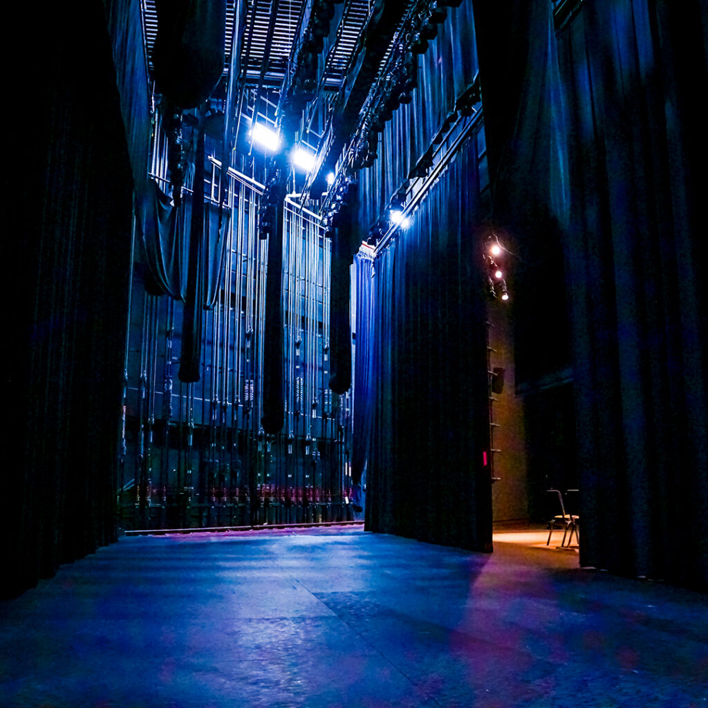 Backstage view of a technical theater, featuring black curtains, bright overhead lights, and a polished floor, with red audience seating in the background.