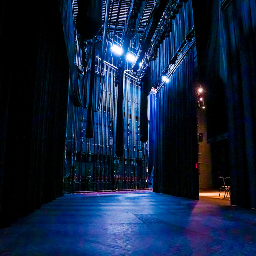Backstage view of a technical theater, featuring black curtains, bright overhead lights, and a polished floor, with red audience seating in the background.
