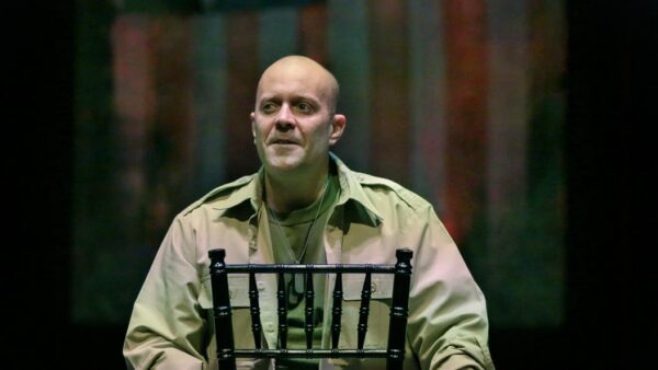 A man in a khaki military shirt sits on a black chair, looking serious against a dark and colorful background.