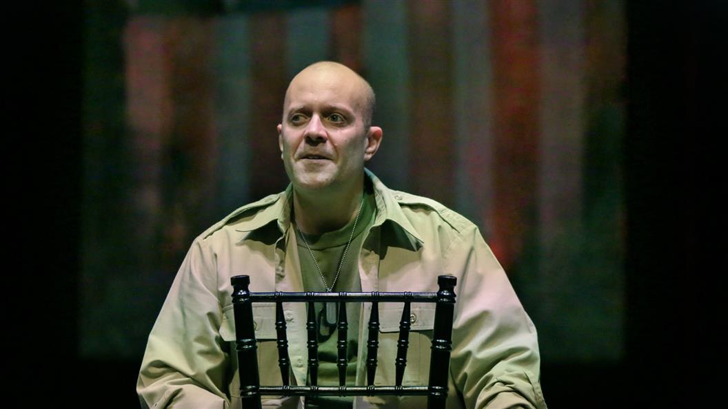 A man in a khaki military shirt sits on a black chair, looking serious against a dark and colorful background.
