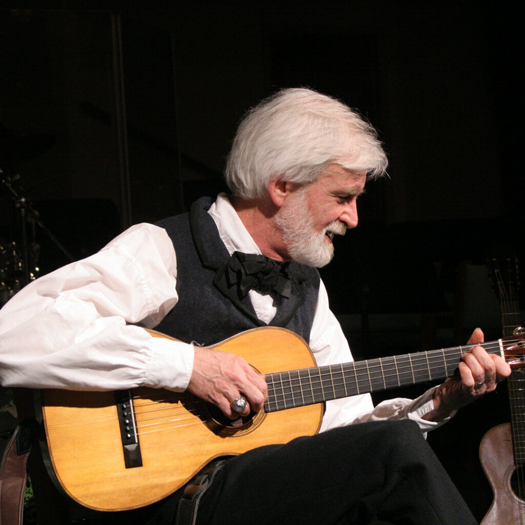 White-haired, bearded man in a vest and bow tie playing an acoustic guitar onstage.