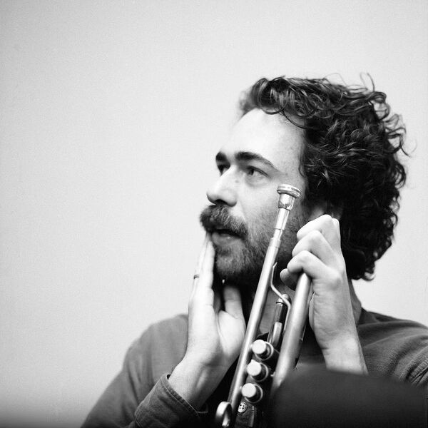 Black-and-white portrait of a bearded man holding a trumpet beside his face while looking left.