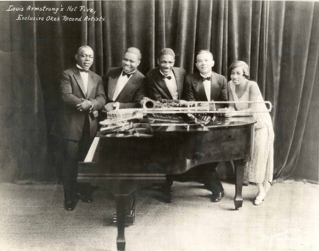 Black-and-white studio portrait of five musicians posed behind a grand piano, with a trombone and other instruments on top.