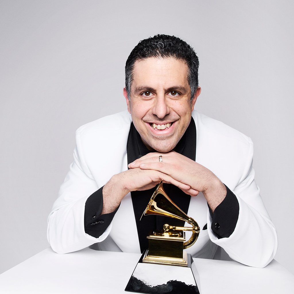 Smiling man in a white jacket leaning over a gold gramophone-shaped trophy on a table.