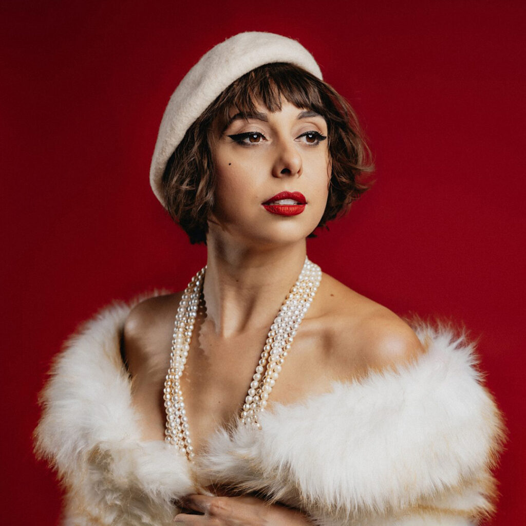 Portrait of a person in a cream beret, layered pearls, and a white faux-fur wrap against a deep-red background.