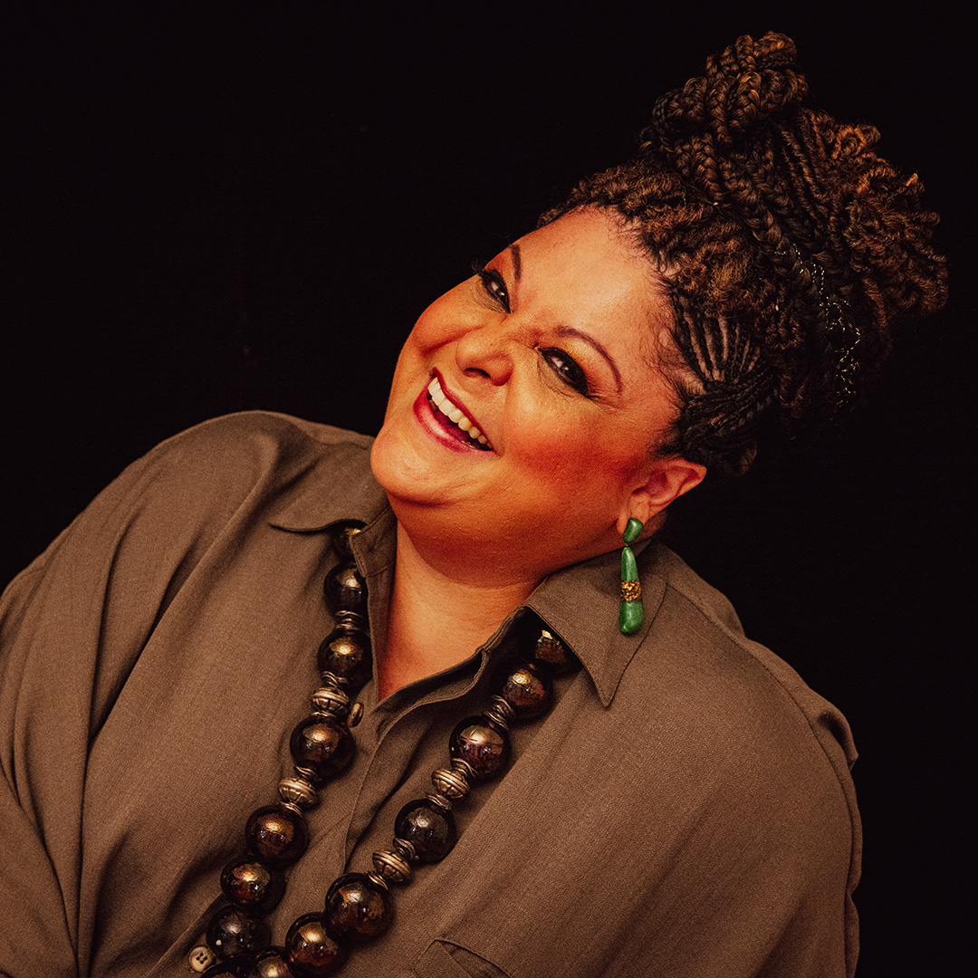 Smiling woman with braided hair, green earrings, and a large beaded necklace posed against a black background.